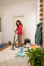 Woman in a red sweater vacuums a patterned rug in a bright entryway with a canister vacuum nearby.