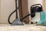 Close-up of a vacuum cleaner with a long hose and a dusty cleaning head on a light carpet, with teal polka-dot rain boots nearby.