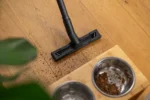 Vacuum cleaner head sucking up crumbs on a wooden floor near two metal dog bowls with dry kibble scattered nearby