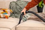 Person vacuuming a beige couch with a handheld hose attachment indoors