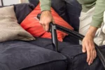 Person uses a handheld vacuum on a dark blue sofa, focusing on the cushions with a flexible hose attachment next to the armrest.