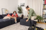 Woman on a dark sofa reads a magazine while a man vacuum-cleans the sofa from a raised ottoman in a cozy living room.