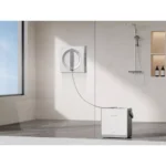 Compact white home spa device on the floor connected by hose to a wall-mounted circular control in a modern bathroom/shower area