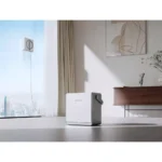 White cube-shaped home appliance on the floor in a modern living room, with a cord running to a wall outlet and a city view through a large window behind it.