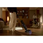 Person vacuuming a hardwood floor in a living room while a caregiver and child play on a blanket in the background in warm lighting.