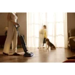 Person vacuuming a living room while a child and a dog stand by sunlit curtains, watching.