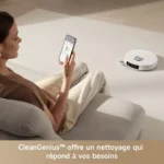 Woman sitting on a sofa using a smartphone while a white robotic vacuum sits on the beige carpet nearby in a living room.