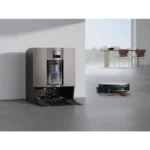 Modern stainless steel coffee machine built into a wall with an open tray, in a bright office space; a robotic vacuum cleaner operates nearby.