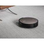 Circular robot vacuum cleaner traversing a light gray carpet near a brown chair leg and metal support