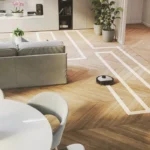 Robot vacuum navigating taped boundary lines on a light wood floor in a modern living room with a gray sofa and plants nearby.