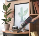 Person using a handheld vacuum to clean a round wooden table with a potted plant, camera, and framed cactus artwork nearby