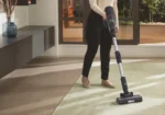 Person wearing a beige sweater vacuuming a light green rug with a cordless stick vacuum inside a modern living room.
