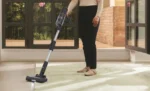 Person vacuuming a light tiled floor with a cordless stick vacuum near an open glass door/entrance area in a bright room.
