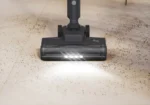 Stick vacuum head on a beige tile floor, sweeping up scattered dirt with its lights on