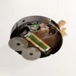 Interior of a circular robot vacuum with exposed brushes and green roller, showing two gray fuzzy wheels and a brown liquid spill inside the chassis.