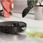 Black robotic vacuum on a light floor projecting a green mapping grid onto the surface ahead as it cleans near a houseplant.