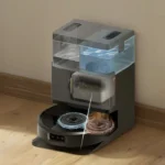 A compact home coffee roaster with a transparent hopper and rotating roasting drums, sitting on a wooden floor.