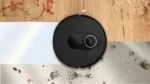 Circular black robot vacuum cleaner moving across a seam between wood flooring and a light gray surface.