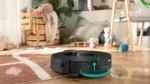 Round black robotic vacuum cleaner on a wooden floor, scrubbing a rug in a child’s playroom filled with toys and crates in the background.