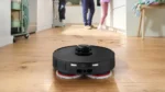 Robotic floor cleaner vacuuming a wooden kitchen floor while two people stand in the background.