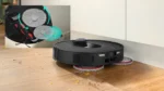 Robotic vacuum on a wooden floor sweeping debris, with side brushes spinning; inset diagram highlights dual rotating brushes with arrows meaning cleaning action