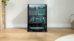 Black Bosch appliance with teal holographic diagrams showing 70°C, placed on a wooden floor in a bright room framed by a plant and basket.
