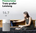 Woman sits on a rug, reaching toward a gray IDEAL smart speaker on a wooden shelf, highlighting its whisper-quiet 16.7 dB performance.
