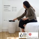 Woman in a sweater sits on a bed beside a white cylindrical Bosch air purifier; dimension labels show 500 mm height and 291 mm width/depth, with award logos in the corner.