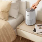 White Philips air purifier on a light wood nightstand beside a bed with beige pillows; a finger pressing the top control.