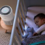 Baby asleep in a crib with a pacifier and stuffed rabbit, beside a white bedside fan on a small table.