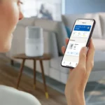 Person holds a smartphone displaying a home automation app in a living room.