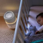 Baby sleeping in a white crib beside a small circular space heater glowing softly. The baby has a pacifier and a blue stuffed animal nearby.
