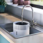 White cylindrical water purifier in a stainless steel kitchen sink with water running from a curved chrome faucet into it, front view.