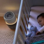 Baby sleeping in a white crib with a modern cylindrical heater on the wooden floor nearby.
