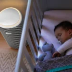 Baby sleeping in a crib with a pacifier, a blue plush toy, and a textured blanket; a round striped Philips device sits nearby on the floor.