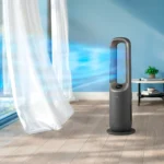 Grey vertical air purifier on a wooden floor beside a window with sheer white curtains, blue light glow in the background, plant and table visible nearby.
