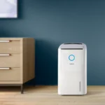 White Philips dehumidifier with a blue circular indicator, standing on a wooden floor beside a dresser in a blue-walled room.
