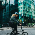 A woman riding a folding bike in an urban street, wearing a helmet and a dark jacket, with glass office buildings in the background.
