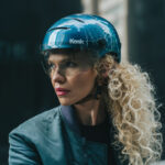 Woman wearing a blue helmet with a visor, looking to the left against a dark background.