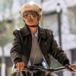 Person wearing a tan helmet and goggles rides a bicycle in an urban setting, wearing a black leather jacket and grey sweater.
