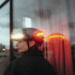 Side profile of a person wearing a white bike helmet with integrated front light, looking left, beside a glass wall with reflections
