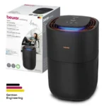 Beurer wellbeing air humidifier in matte black, cylindrical with a blue illuminated control ring, shown beside its packaging box.
