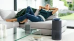 Woman lounging on a light gray sofa in a bright living room, head resting back, with a round speaker on the glass coffee table and a tall cylindrical speaker nearby.