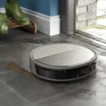 Robot vacuum (Samsung) sits on a tiled floor near a bench with green rain boots, by a glass door leading outside.
