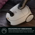 White canister vacuum with a foot pressed on the on/off control, highlighting ergonomic foot control feature (German caption: 'ERGONOMISCHE FUSSBEDIENUNG')