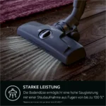 Close-up of a vacuum cleaner floor head on a wooden floor with scattered dust; a dark banner at the bottom displays the German text 'STARKE LEISTUNG' (strong performance).