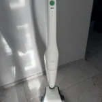 White cordless stick vacuum standing upright on a tiled floor next to a white wall. The green power button is visible on the handle.