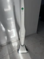White cordless stick vacuum standing upright on a tiled floor next to a white wall. The green power button is visible on the handle.