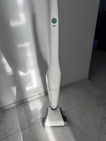 White cordless stick vacuum standing upright on a tiled floor next to a white wall. The green power button is visible on the handle.