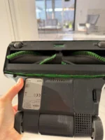Hand holding the underside of a vacuum cleaning head with two green rotating brushes exposed.